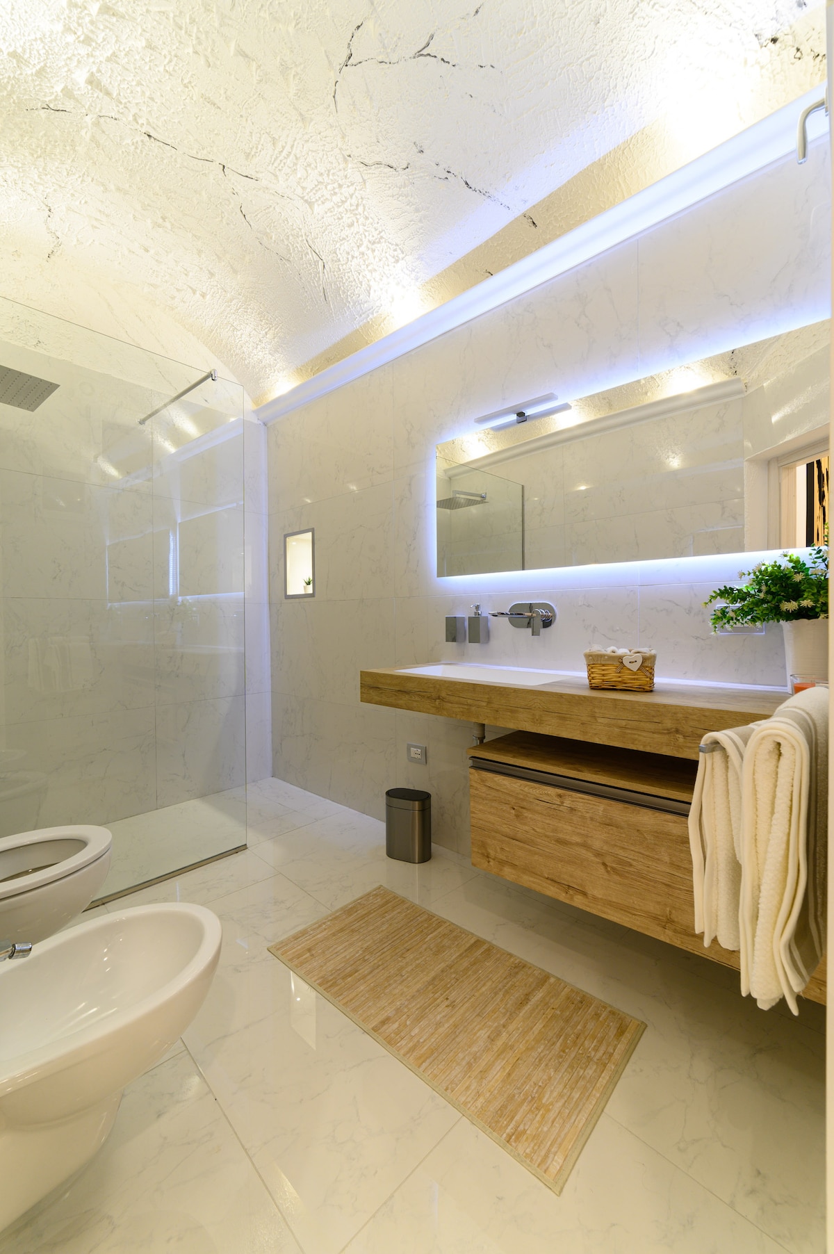 The modern bathroom features a spacious walk-in shower with clear glass and stylish fixtures. A wooden vanity holds neatly organized toiletries, while a soft rug lies on the marble floor. Ambient lighting highlights the sleek design, creating a clean and inviting environment.