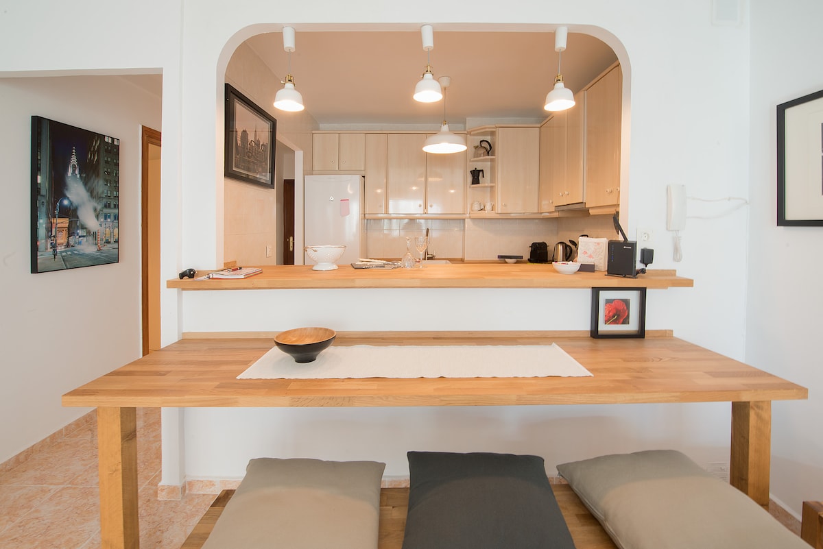 A wooden table with a light-colored runner is positioned in front of an open kitchen, complemented by soft cushions underneath for seating. Shelves above display simple decor items, while bright overhead lights enhance the welcoming ambiance.