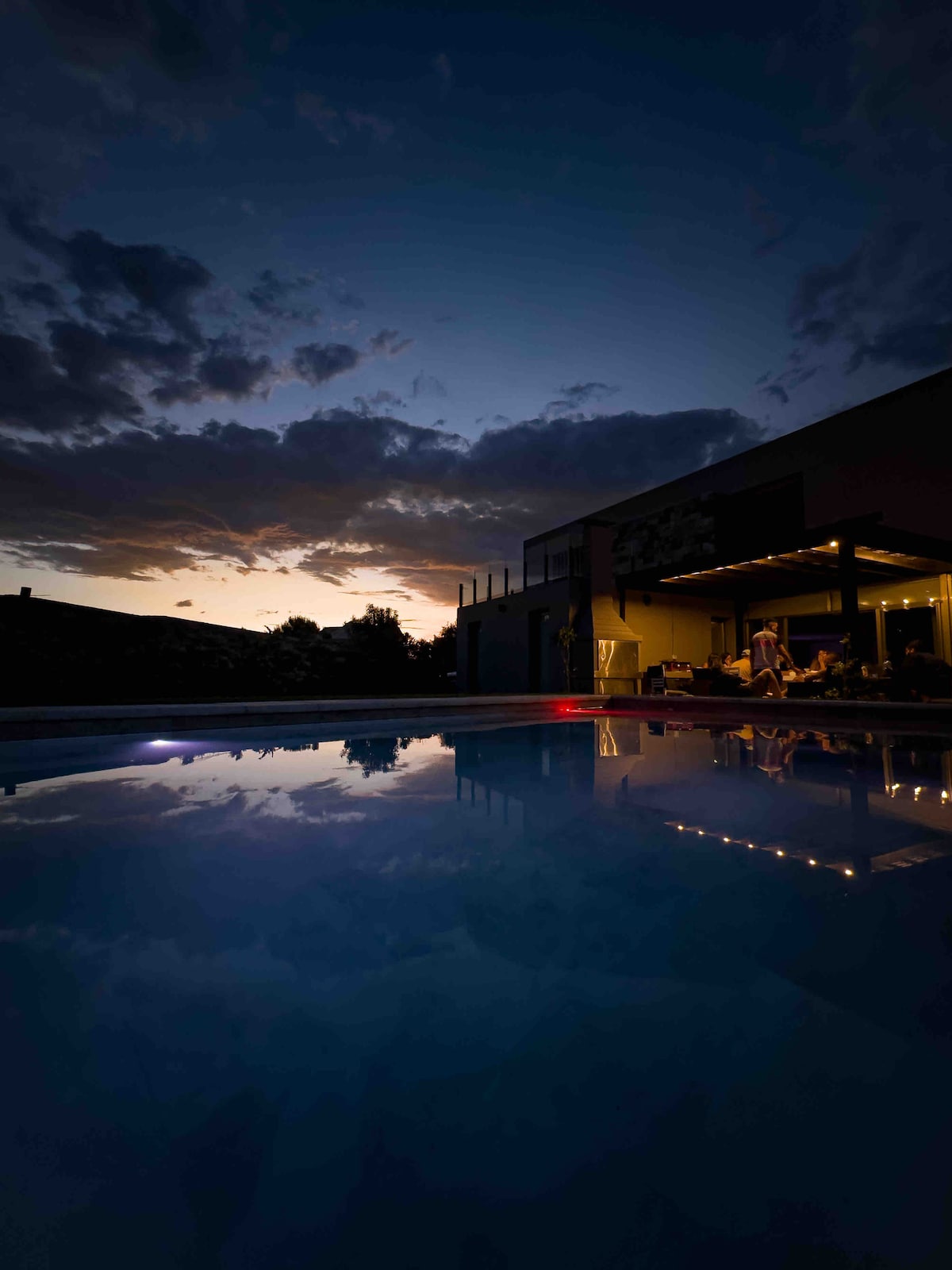 An inviting outdoor space is shown with a serene swimming pool reflecting the colorful sky at dusk. Warm light illuminates a shaded seating area, where people are gathered, creating a cozy atmosphere as the sun sets behind the house.