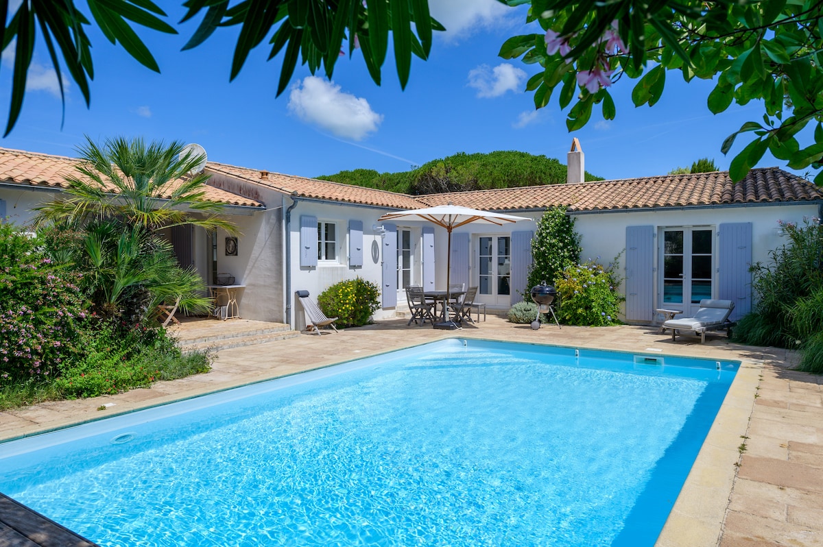 A spacious outdoor area is depicted, showcasing a large swimming pool surrounded by a sunbathing deck. Sun loungers are positioned near the pool, and a shaded dining area with an umbrella provides seating. Lush greenery and bright blue skies enhance the ambiance.