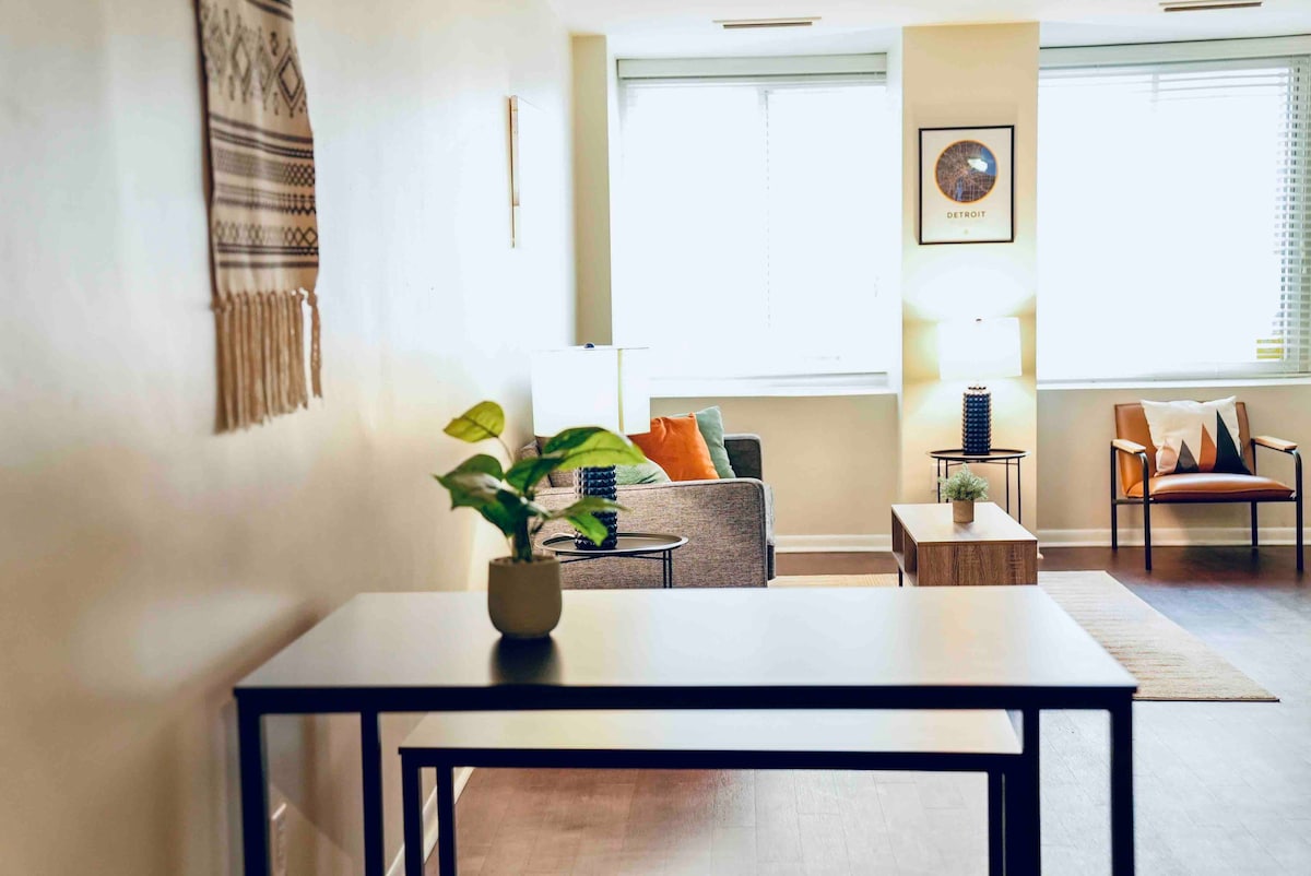 A welcoming living area features a small dining table with a potted plant, surrounded by a cozy sofa and armchair. Natural light filters through the large windows, which are adorned with simple blinds, creating a bright and casual atmosphere.