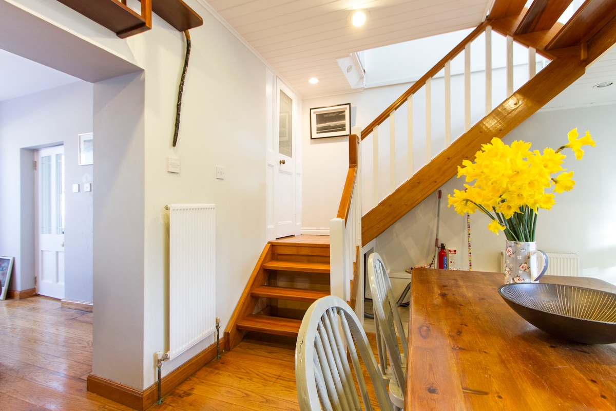 A welcoming entryway features a wooden staircase leading to the upper level. A simple table is adorned with a vase of bright yellow flowers. Light floods in from a nearby window, enhancing the neutral tones of the walls and wood flooring.
