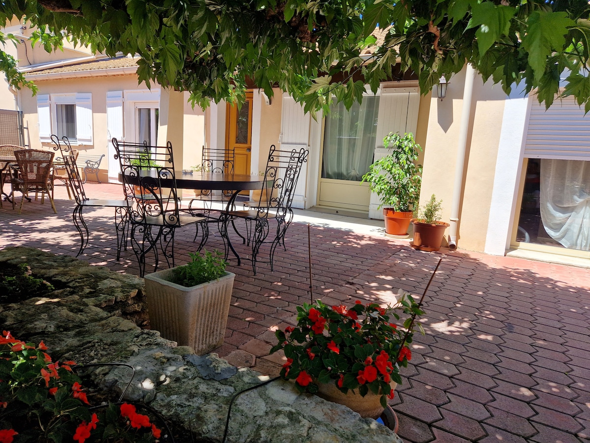 An outdoor terrace is shaded by lush greenery, featuring a large table surrounded by metal chairs. Potted plants with vibrant flowers add color to the space, which is set against a backdrop of a light-colored facade with large windows.