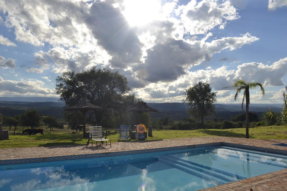 A swimming pool is surrounded by lush green grass, with sun loungers positioned nearby. The sky is filled with fluffy clouds, and distant hills are visible in the background. Shade is provided by palm trees and thatched umbrellas, creating inviting outdoor spaces.