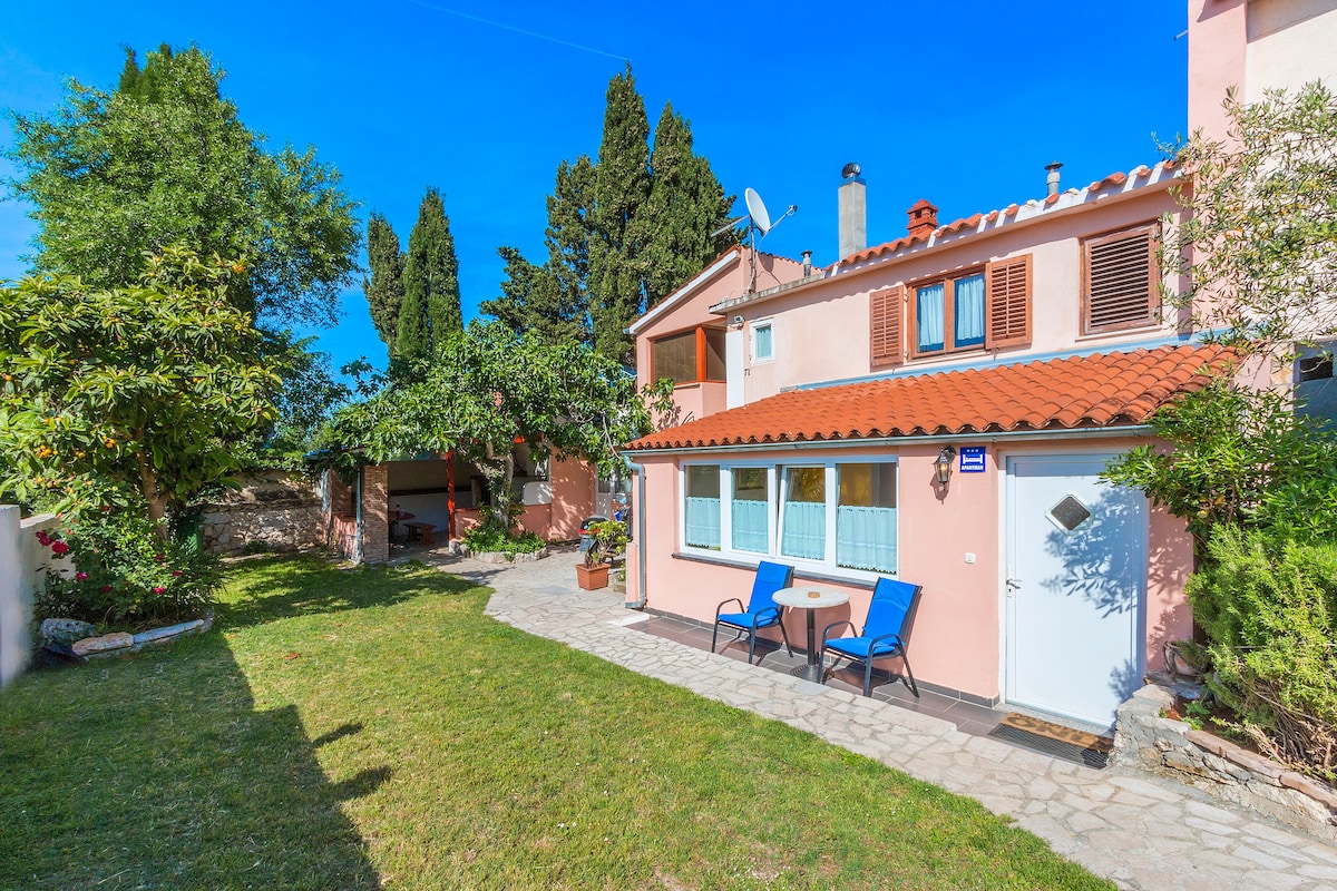 The exterior of the apartment features a well-maintained yard with green grass and decorative trees. A small patio area is visible with a table and two chairs. The building has a charming pink facade complemented by a tiled roof and various window designs.