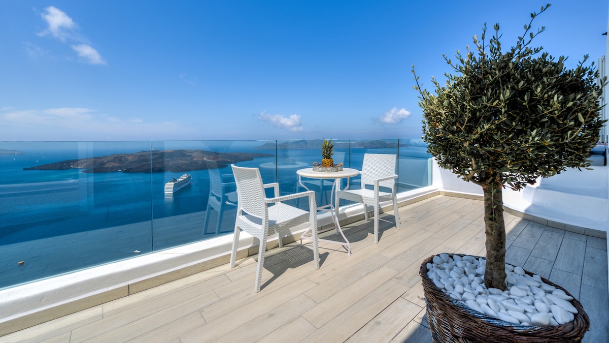 A spacious terrace overlooks a serene body of water, featuring a glass railing that enhances the view. Two white chairs and a small table are arranged for relaxation, alongside a decorative plant in a woven basket filled with white stones.