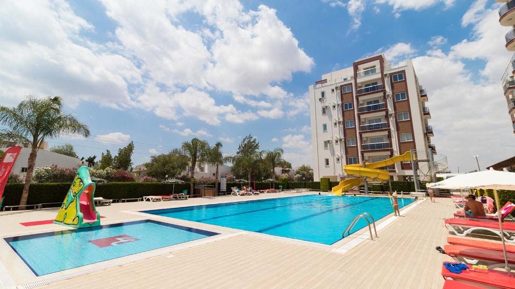 A spacious swimming pool is featured, accompanied by a playful slide and lounge chairs placed around the pool area. Tropical palm trees are visible in the background, and a modern multi-story building stands adjacent, providing a relaxed atmosphere.