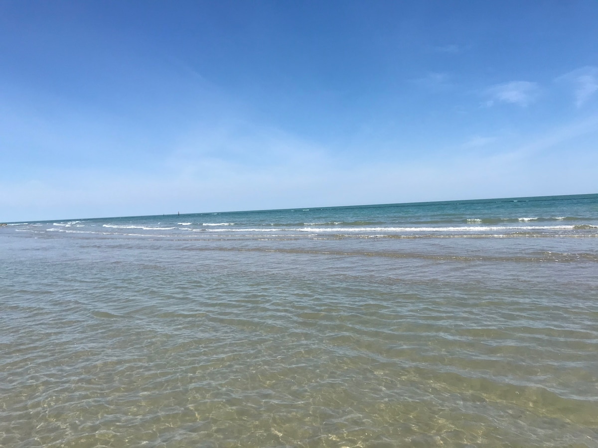 The image captures a serene beach scene with gentle waves lapping against the shoreline. The clear, shallow water extends toward the horizon, where the sea meets a bright blue sky, creating a sense of tranquility and openness.