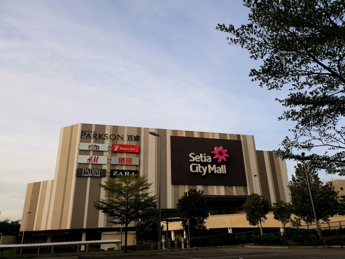 The exterior of Setia City Mall is presented, showcasing a modern architectural design with distinctive striped panels. Store signage, including well-known brands, is visible. Lush greenery is seen in the foreground, enhancing the urban landscape. The sky exhibits a soft gradient, indicating early morning or late afternoon.