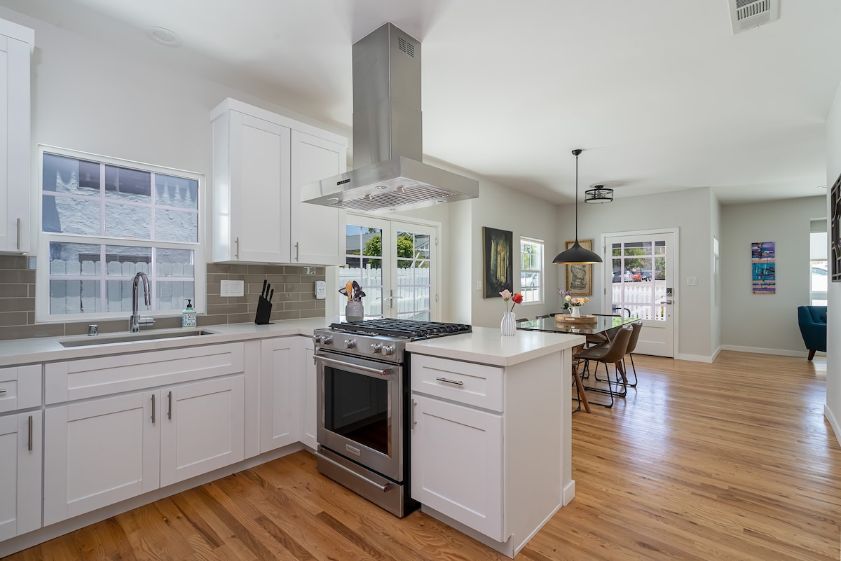 The modern kitchen features stainless steel appliances, including a large gas stove and a hooded vent. White cabinetry and countertops create a bright atmosphere, while hardwood flooring enhances the open layout. Natural light enters through multiple windows, offering views of the outdoor space.