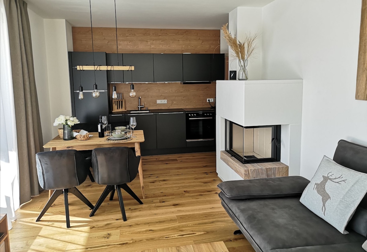 An open kitchen and dining area are showcased, featuring sleek black cabinetry and wooden accents. A dining table with two upholstered chairs is set for a meal. A contemporary fireplace is positioned nearby, enhancing the modern decor with a touch of warmth.