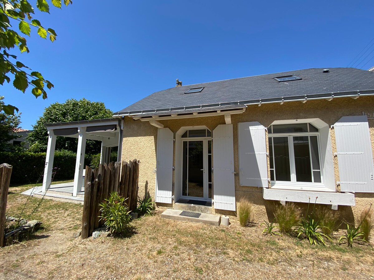 A charming house from the 1930s is displayed, featuring white window shutters and a welcoming entrance. The exterior showcases a blend of light tones, lush greenery, and a clear blue sky, with a patio area visible to the side. The yard is surrounded by a natural wood fence.