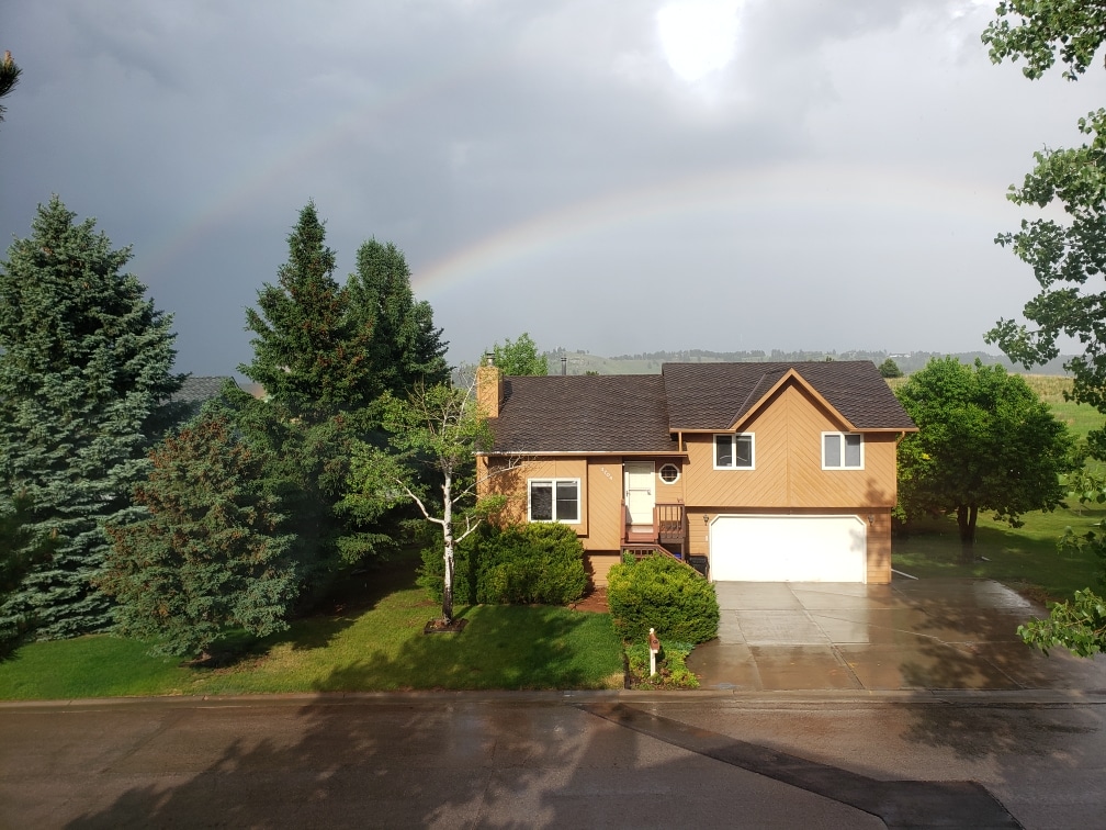 A welcoming home is set in a lush landscape, surrounded by tall trees. A vibrant rainbow arcs over the house, contrasting with the gray sky. The driveway leads to a two-car garage, while the well-maintained yard offers a green backdrop.