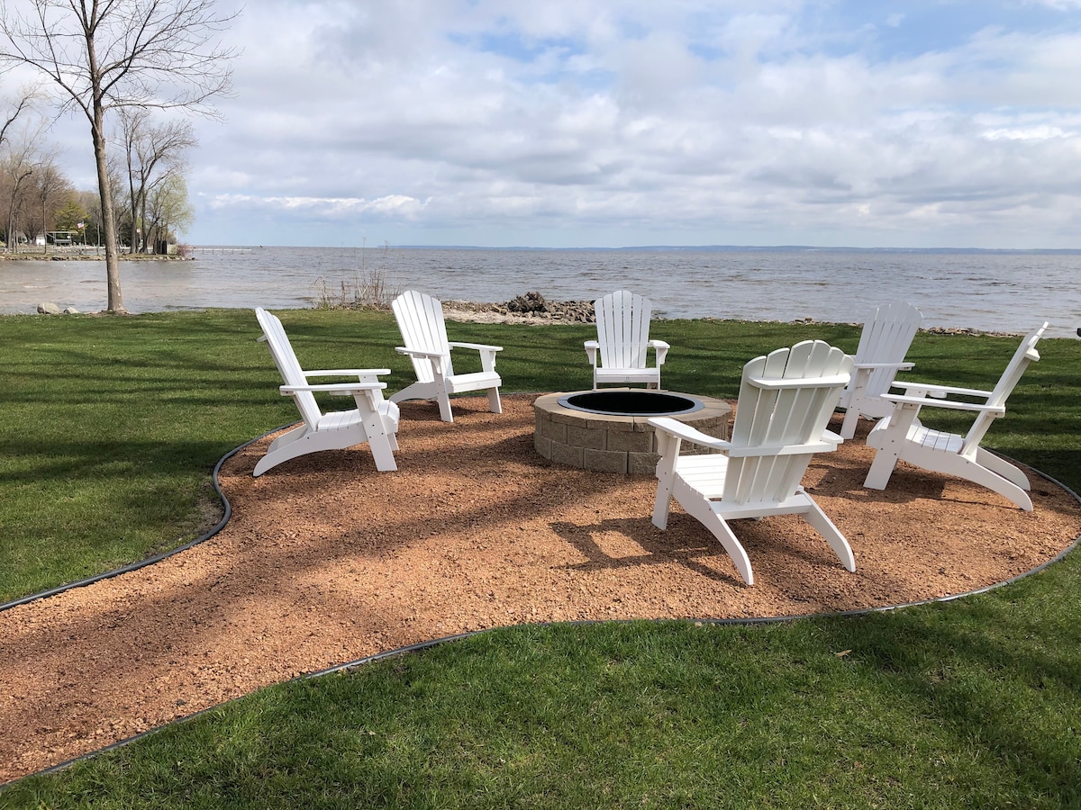 A firepit surrounded by five white Adirondack chairs is situated on a sandy area, overlooking the lake. Green grass frames the space, with trees dotting the shoreline under a partially cloudy sky.