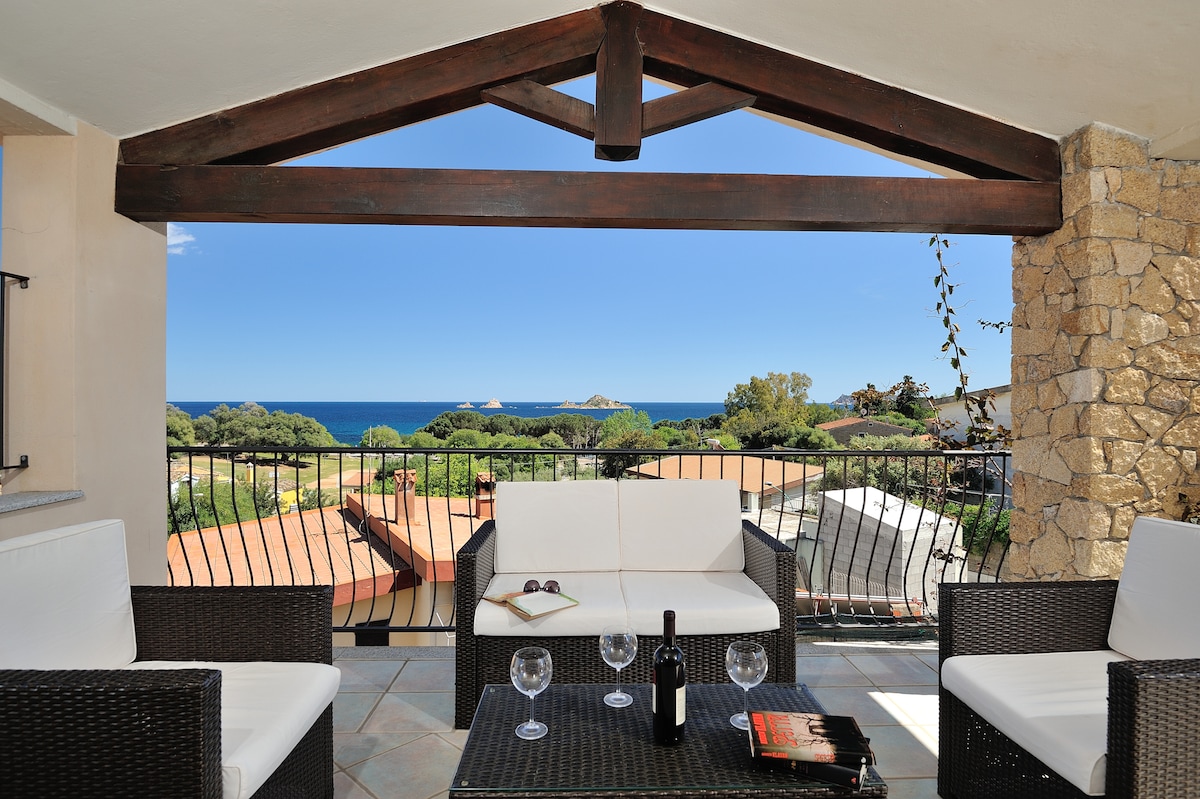 A balcony is featured with a wooden beam ceiling, showcasing a seating area with a dark wicker sofa and table. Scenic views of the sea and surrounding landscape are visible beyond the railing, enhancing the outdoor space for relaxation.