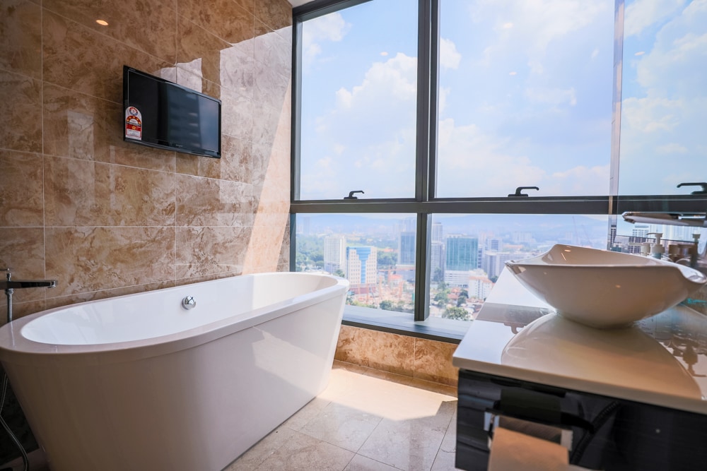 A modern bathroom features a freestanding bathtub positioned near a large window, offering a panoramic view of the cityscape. A sleek countertop with a pedestal sink and a wall-mounted television are visible, alongside elegant tiles that enhance the space's contemporary design.