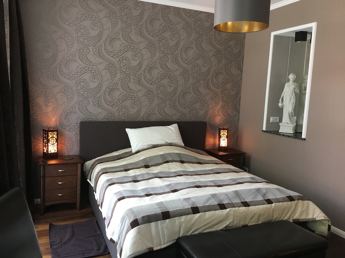 A cozy sleeping area features a spacious bed with a neutral-colored striped blanket. Bedside tables with decorative lamps provide soft lighting. A wall-mounted mirror reflects a statue, and subtle wallpaper adds texture to the room, complemented by warm wooden flooring.