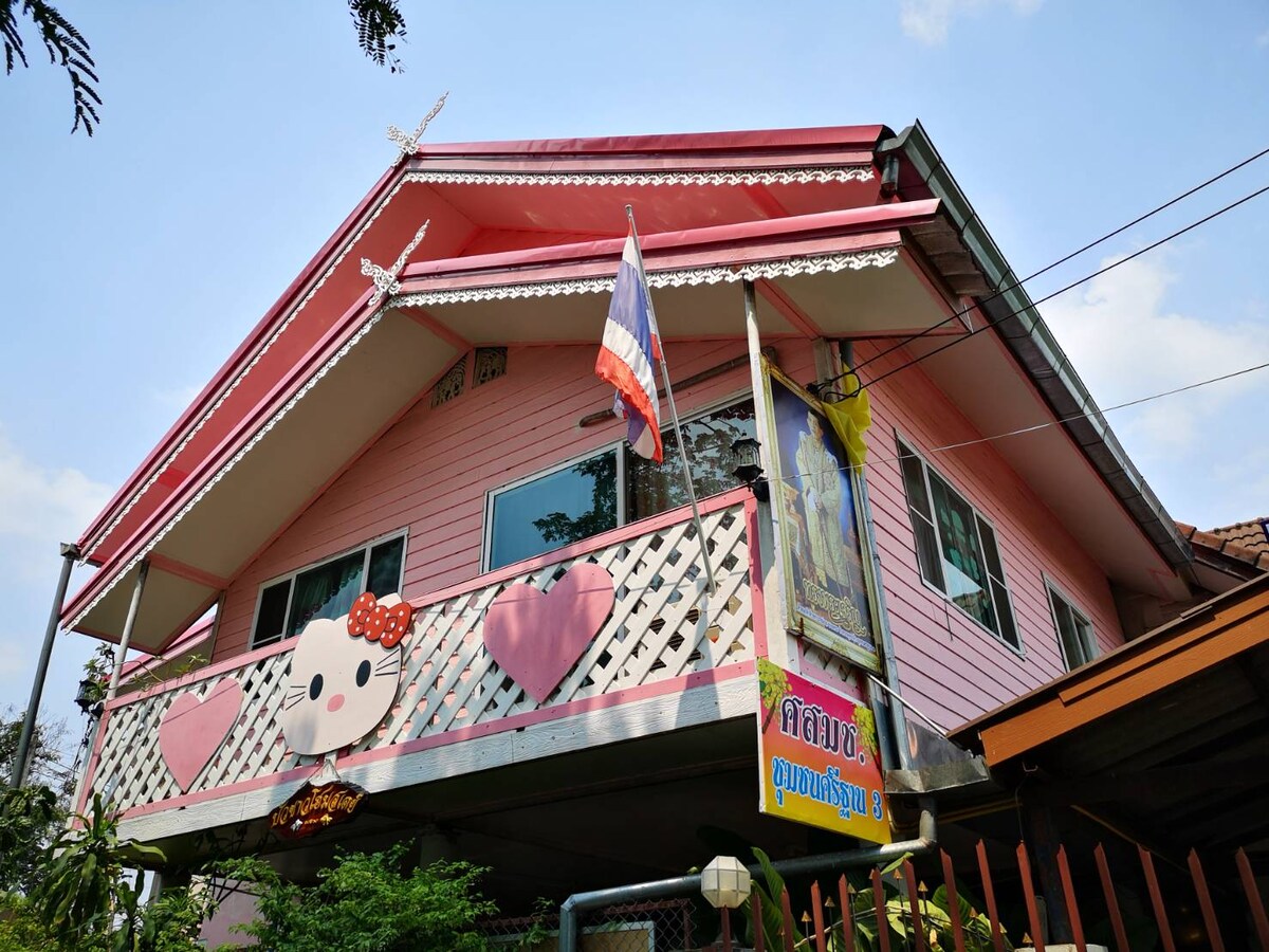 A two-story house features a playful pink exterior with heart-shaped decorations and a large balcony. Flags are displayed in front, surrounded by greenery. The roof has a classic design with decorative elements, providing a cheerful charm to the property.