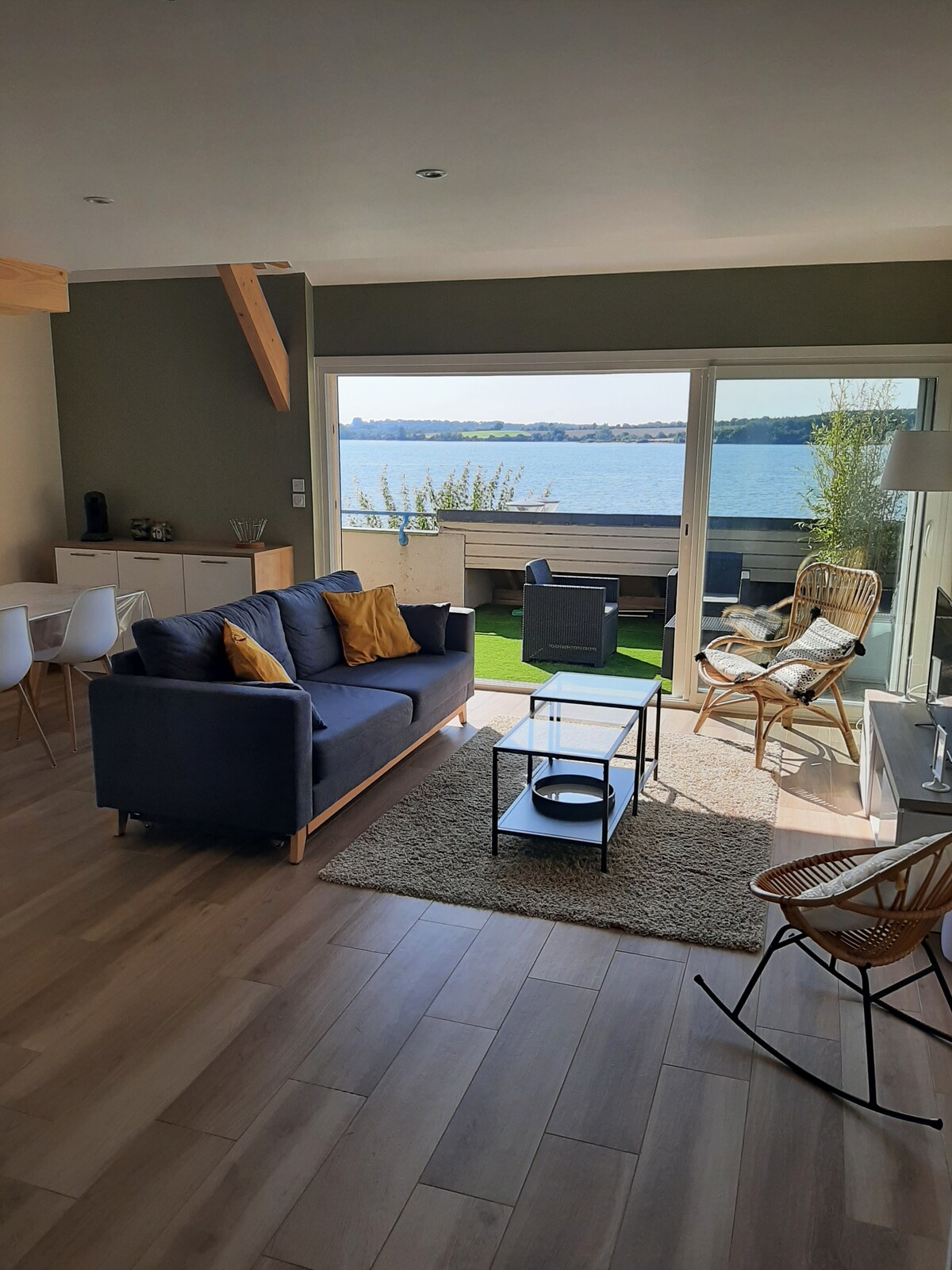 The bright open living area features large glass doors offering unobstructed views of the lake. A comfortable blue sofa is complemented by yellow cushions, surrounded by a glass coffee table and chairs. Natural light fills the space, enhancing the serene atmosphere.
