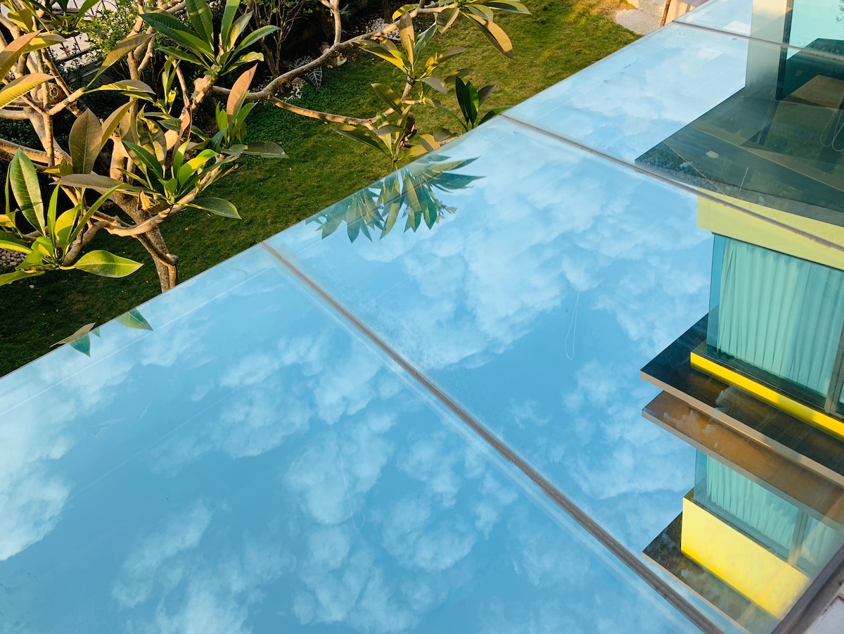 A glass surface reflects a clear blue sky with fluffy clouds, surrounded by lush greenery and trees. The image captures the tranquil outdoor atmosphere, with hints of a building structure visible in the background.