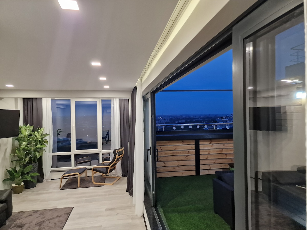 A spacious living area features modern furnishings, including a relaxed chair and a soft area rug. Large glass doors open to a terrace, offering expansive views of the city skyline against an evening sky. Indoor plants add a touch of greenery to the room.