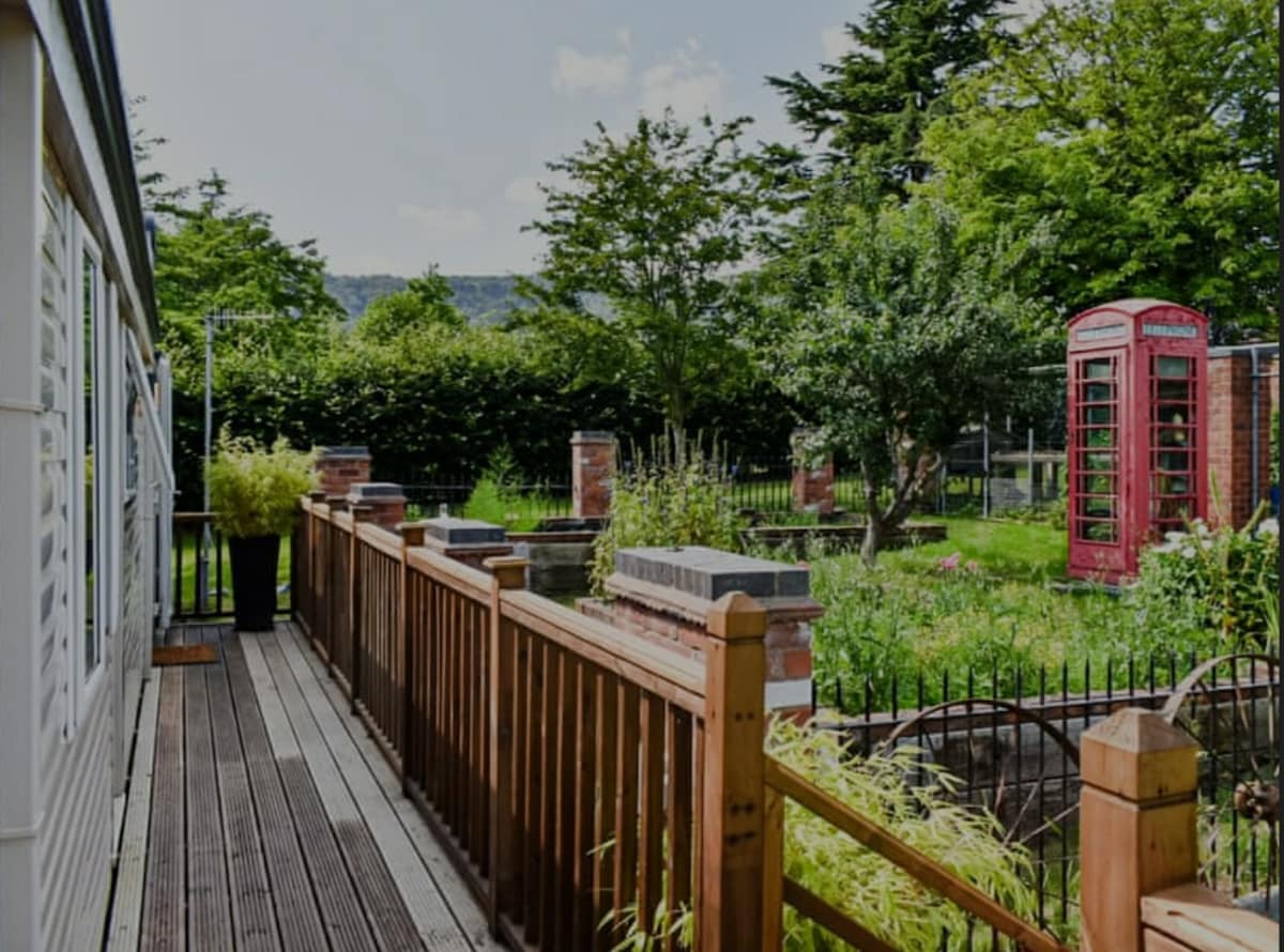 A wooden deck stretches from the lodge, leading towards a landscaped garden featuring lush greenery, flower beds, and a classic red telephone box. Hilltops are visible in the background, creating a serene outdoor environment surrounded by well-maintained landscaping.