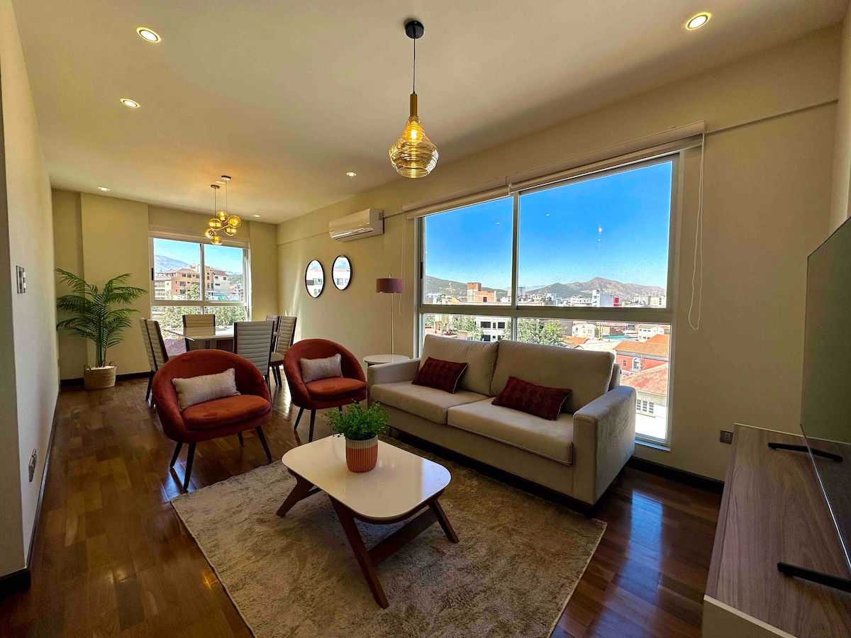 A modern living area is furnished with a plush sofa and two accent chairs arranged around a minimalist coffee table. Light pours in through large windows, revealing a scenic view of the surrounding mountains and cityscape. The space is complemented by tasteful lighting fixtures.