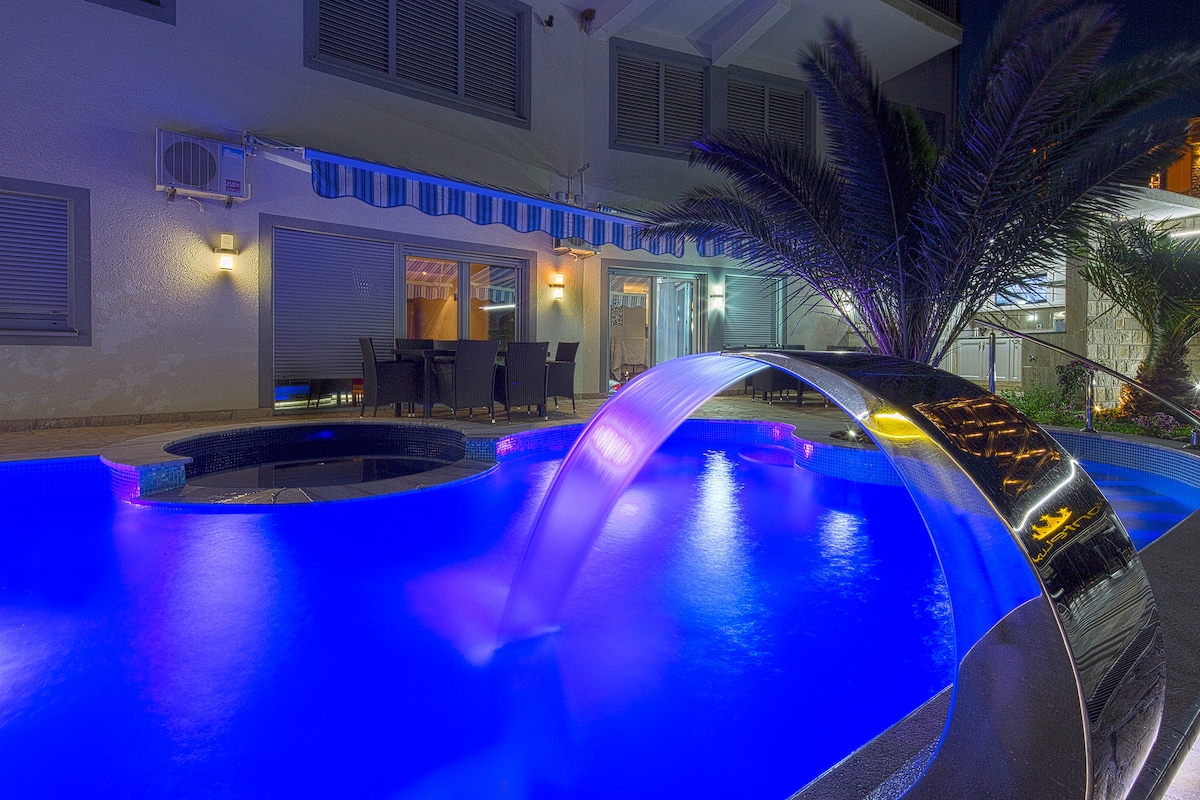 A night view of the swimming pool features illuminated water in shades of blue. A stylish water feature creates a gentle cascade into the pool, while a jacuzzi and lounge area with dining furniture are visible in the background, surrounded by tropical plants.
