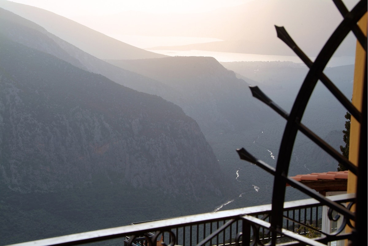 A scenic view is framed by a wrought iron railing, showcasing rolling hills and the distant Corinthian Gulf under a soft haze. The landscape features winding valleys and a river glistening in the sunlight, creating a serene natural backdrop.