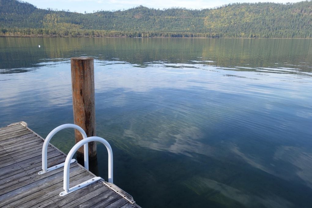 A serene view of Priest Lake showcases the calm waters reflecting the surrounding hills. A wooden dock extends into the lake, featuring a sturdy post and white railing, inviting guests to enjoy moments by the water's edge.