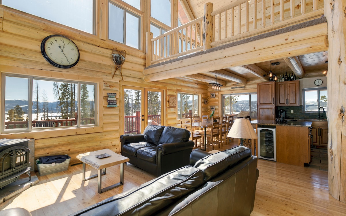 The interior space features high ceilings with large windows providing natural light and stunning mountain views. A cozy living area is furnished with a dark leather sofa and a wooden coffee table. The dining area includes a large wooden table surrounded by chairs, complemented by a well-equipped kitchen.