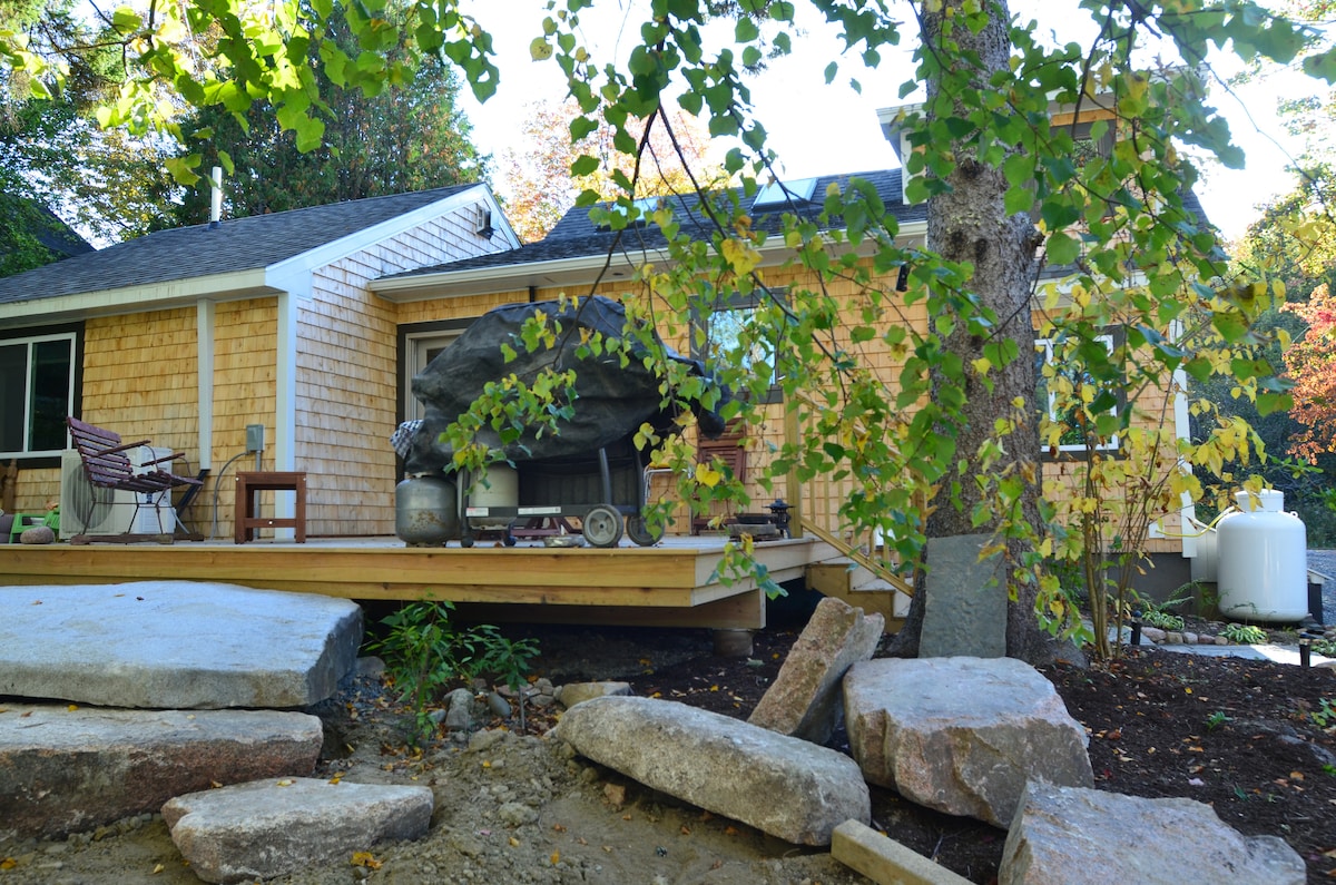 The exterior of the house features natural wood siding and a spacious wooden deck, framed by vibrant green foliage. The entrance is partially covered by a large rock formation, surrounded by natural stone pathways leading to the home.
