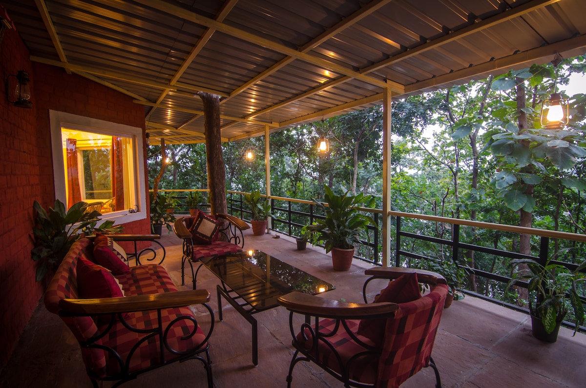 A covered outdoor seating area is featured, offering views of the surrounding greenery. Comfortable seating with cushions is arranged around a central table, while plants adorn the space. Soft lighting from hanging fixtures creates a relaxing ambiance, inviting guests to enjoy the natural surroundings.