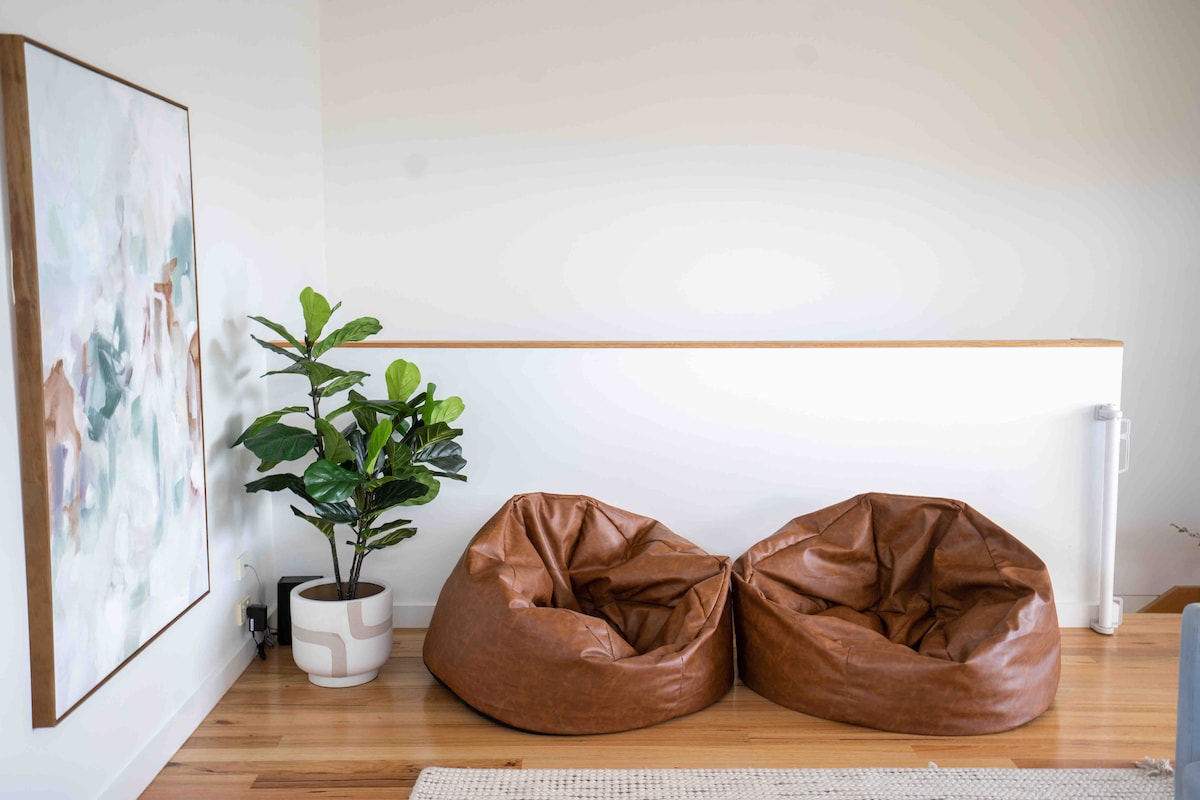 A cozy corner is showcased with two leather bean bags in earthy tones. A decorative plant in a contemporary pot adds a touch of greenery. A light-colored area rug complements the wooden flooring, and an abstract painting hangs on the wall, enhancing the overall aesthetic.