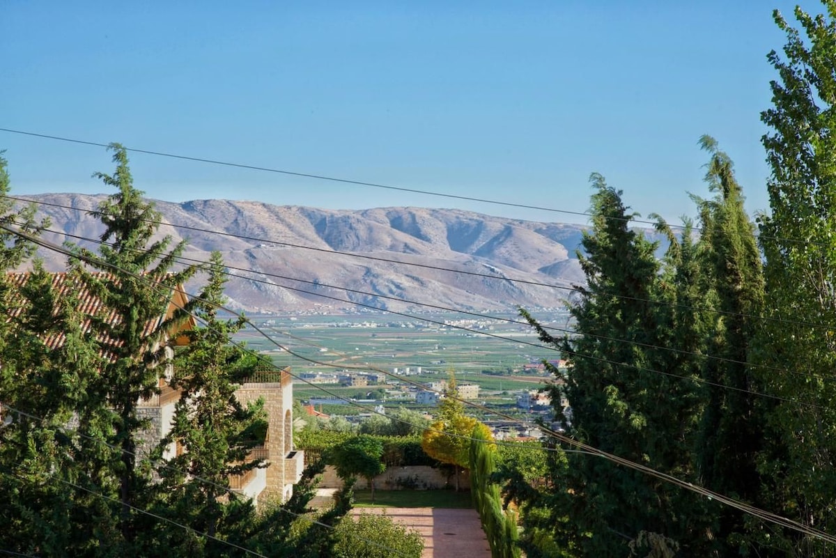 Distant views of rolling hills and a green valley are framed by nearby trees, creating a serene outdoor scene. The landscape is enhanced by clear skies, with subtle outlines of fields and distant mountains visible in the background.