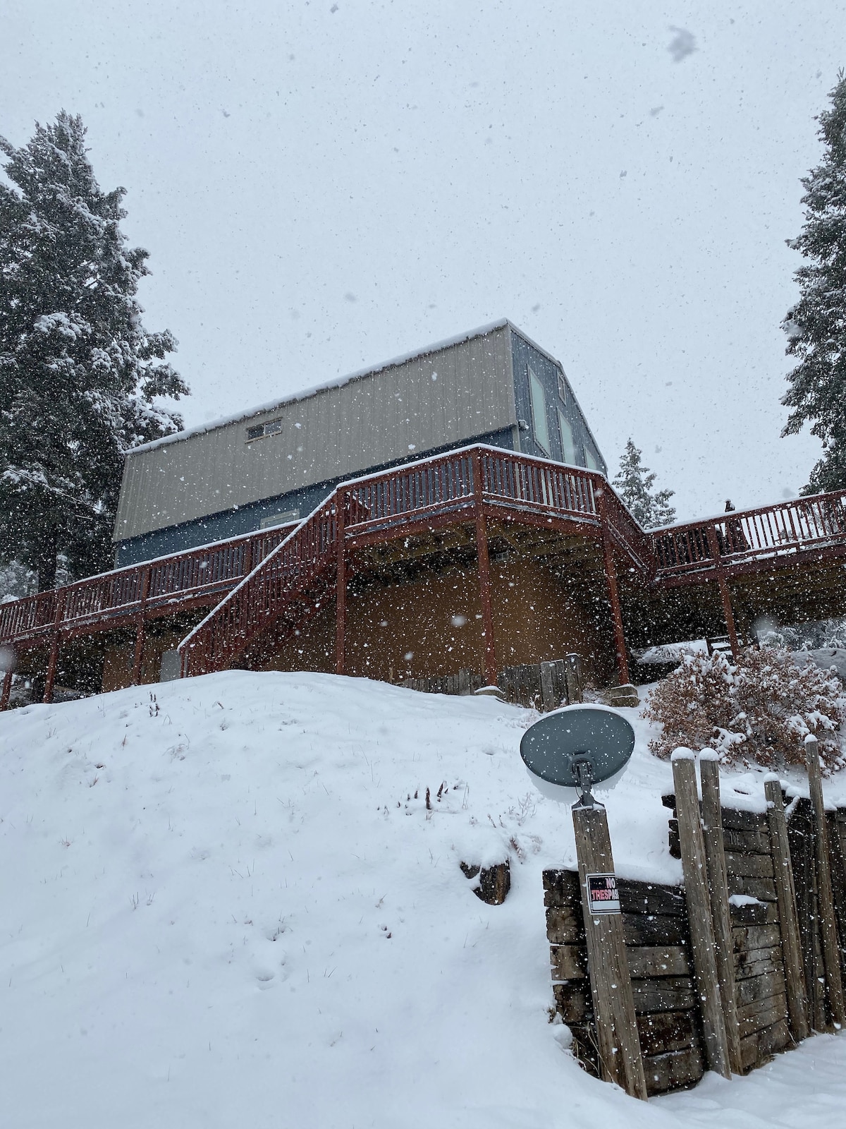 A multi-level house sits surrounded by snow-covered ground, with snowflakes steadily falling. The building features large windows on the upper level, allowing natural light to enter. A wooden deck wraps around the structure, complementing the surrounding winter landscape.