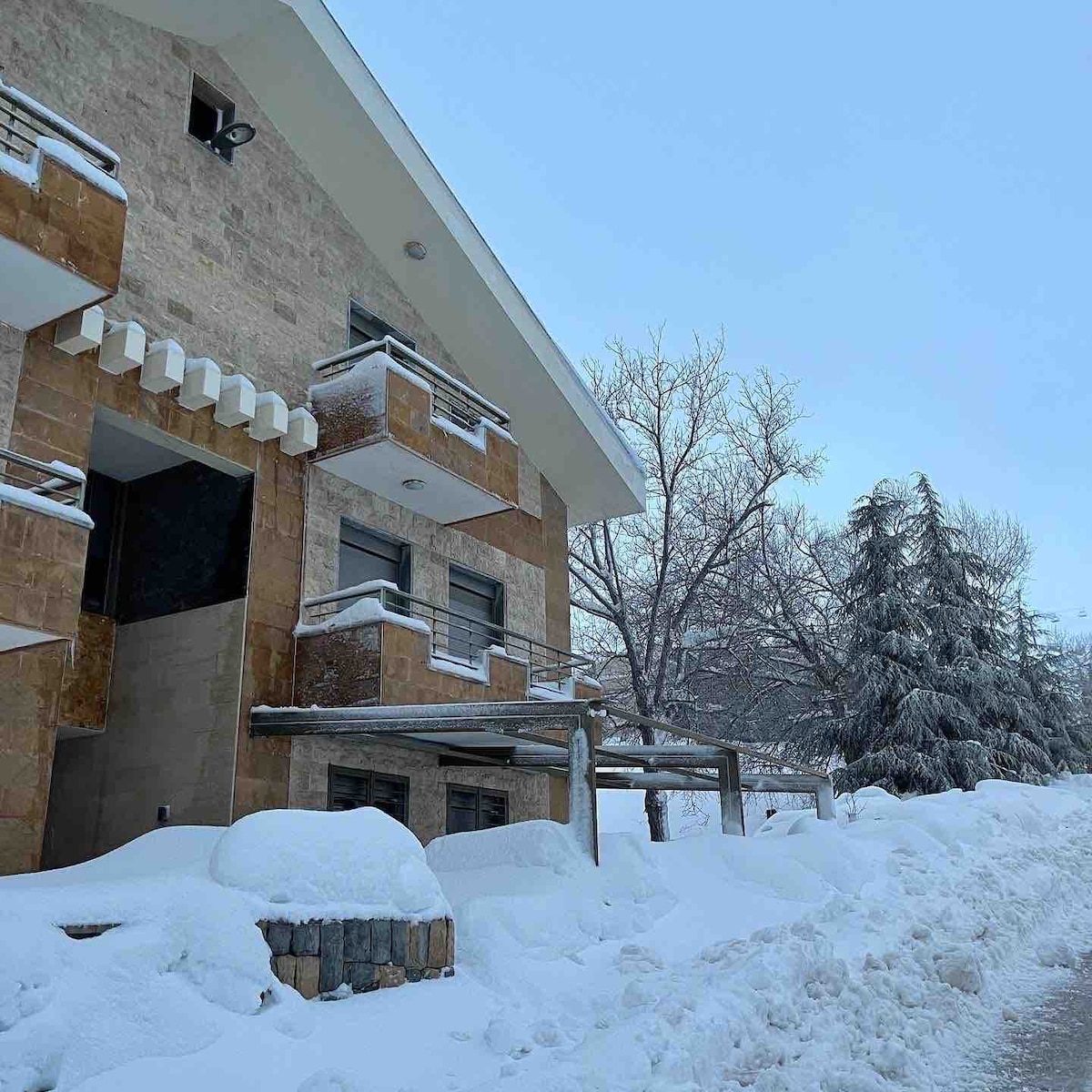 The exterior of the house is partially covered in fresh snow, showcasing a modern stone façade and multiple balconies. Nearby trees are blanketed in white, and the sky is a soft blue, indicating a clear winter day.