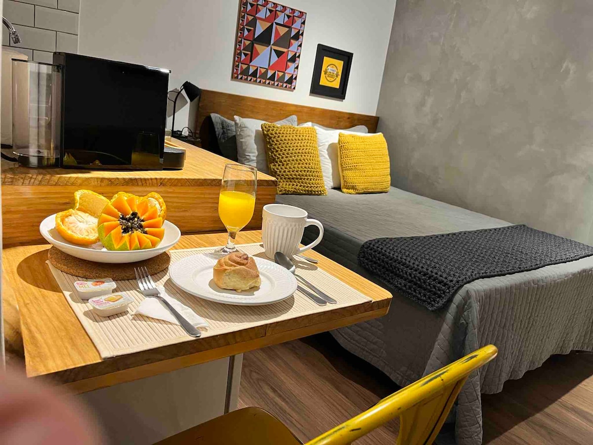 A cozy studio features a spacious bed with textured pillows and a complementary throw. A breakfast setting is displayed on a wooden table, including a fruit plate, a pastry, and a mug. Artwork adorns the wall, and a kitchenette area is visible in the background.
