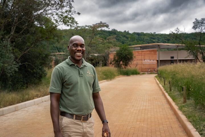 A man wearing a green t-shirt with a Lapalala Wilderness School logo smiles broadly while standing in front of a building called the Lapalala Wilderness School