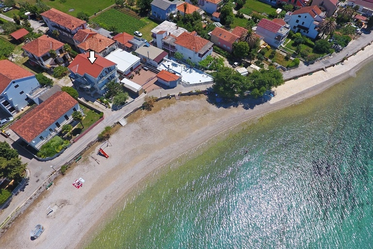 An aerial view showcases the beachfront location of the apartment, surrounded by a residential area. The sandy shore and clear waters are visible, with several buildings lining the street. Green patches of grass and a few trees enhance the neighborhood's landscape.