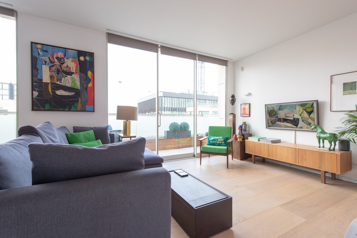 A modern living area features a spacious grey sofa and a wooden coffee table. Bright artworks adorn the walls, and natural light enters through large glass doors, leading to a balcony. A green accent chair and decorative items contribute to the stylish ambiance.