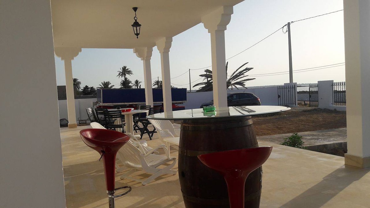 The outdoor terrace features a large round table atop a barrel, surrounded by red and white bar stools. Comfortable lounge chairs are positioned for relaxation, with a view of palm trees and the sea in the background. Natural light enhances the inviting atmosphere.