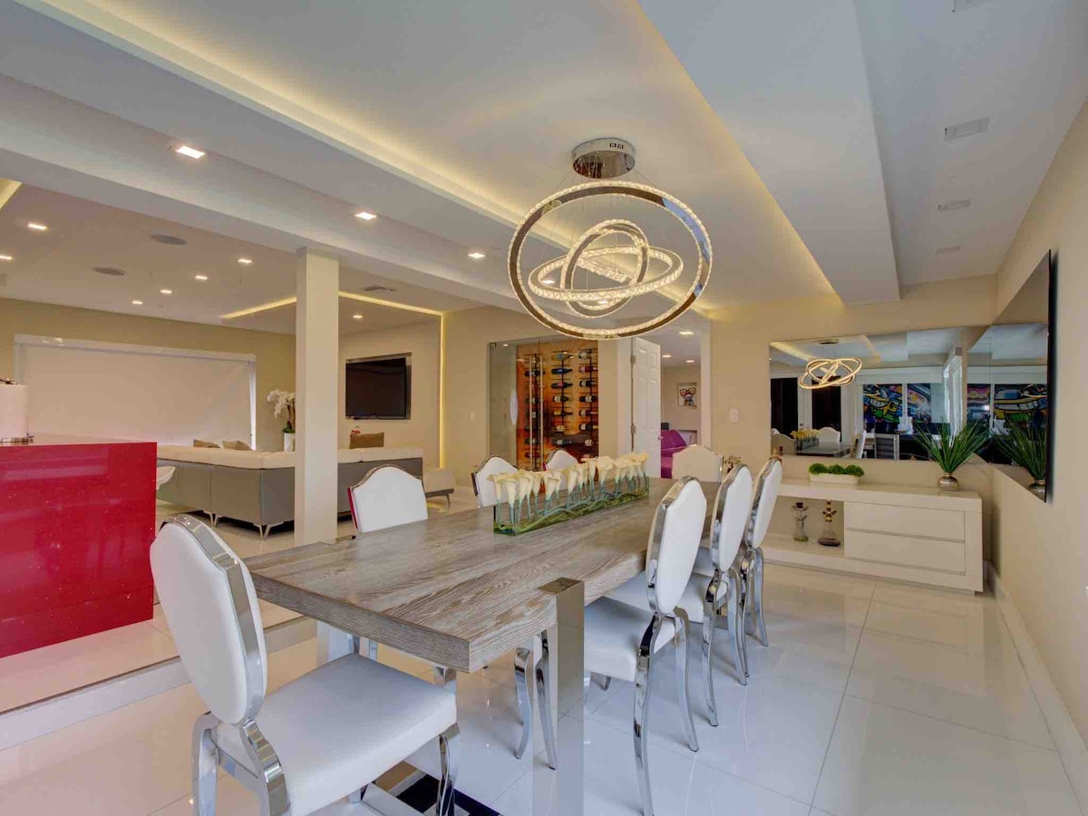 A modern dining area features a sleek wooden table surrounded by elegantly designed chairs. A decorative centerpiece of greenery enhances the setting. Soft lighting emanates from a contemporary chandelier overhead, while large mirrors and a vibrant wine display add visual interest to the spacious open layout.