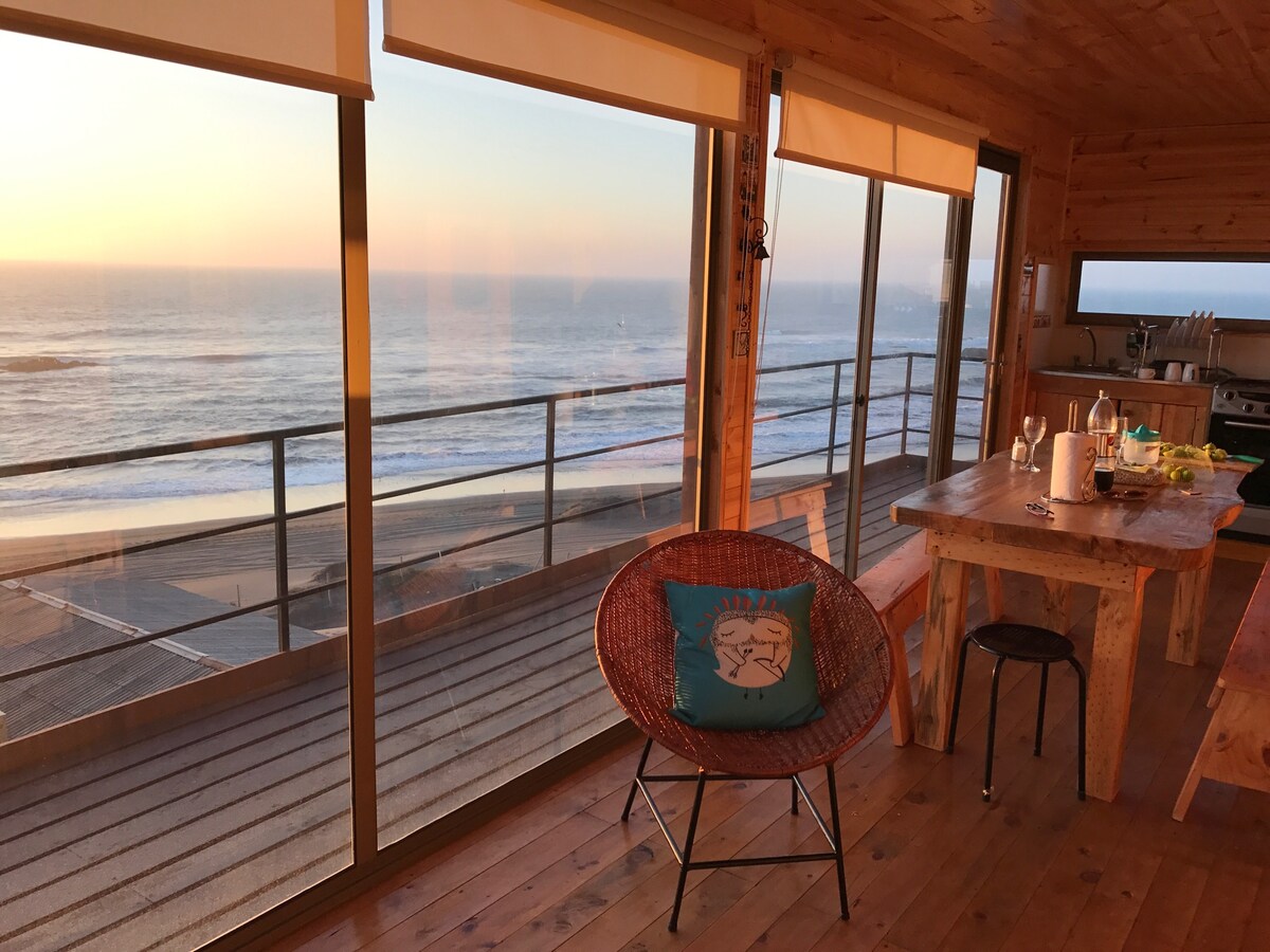 Large windows offer an unobstructed view of the beach and ocean, allowing natural light to fill the room. A wooden table is paired with colorful chairs, while a cozy armchair with a decorative cushion invites relaxation. The relaxed atmosphere is enhanced by warm wooden tones and soft sunlight.
