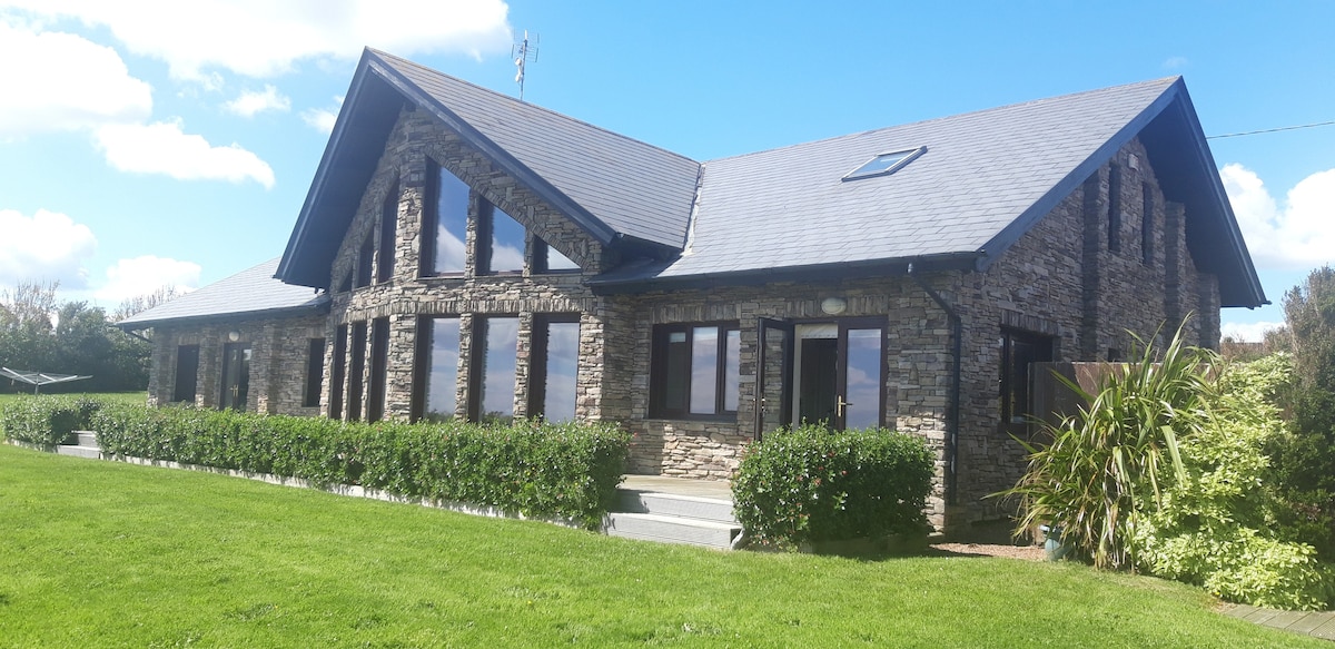 The log cabin features a stone exterior with large windows, allowing natural light to fill the interior. A well-maintained lawn surrounds the property, while a subtle garden adds greenery. The structure exhibits a sloped roof and is set against a clear blue sky.