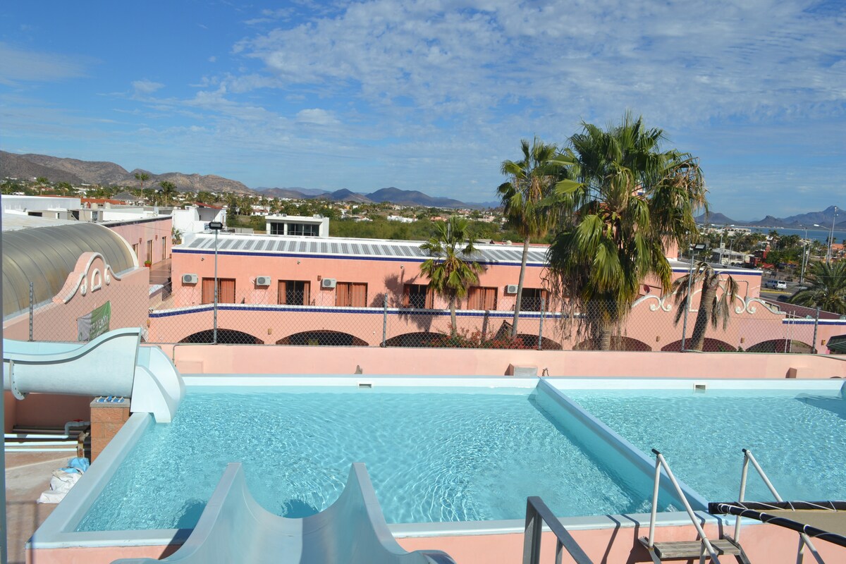 A rooftop pool area features a spacious swimming pool with tranquil blue waters. Palm trees and distant mountains create a scenic backdrop. A water slide is present, adding a playful element to the space, while a sun deck provides an open area for relaxation.
