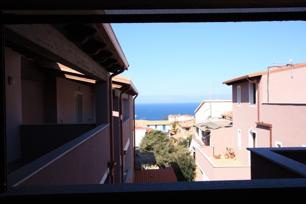 A view through a window frames the serene blue sea, contrasting with the nearby buildings. The exterior offers a blend of natural elements with trees visible below and rooftops of nearby houses. The open sky enhances the view, providing a sense of openness.
