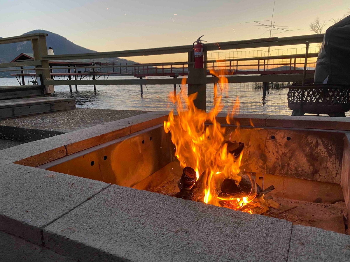 A fire pit is surrounded by stone edging, with flames dancing from the center. The warm glow contrasts with the serene lake view and the silhouette of a mountain in the background. A dock is visible, along with wooden railings that frame the tranquil scene.