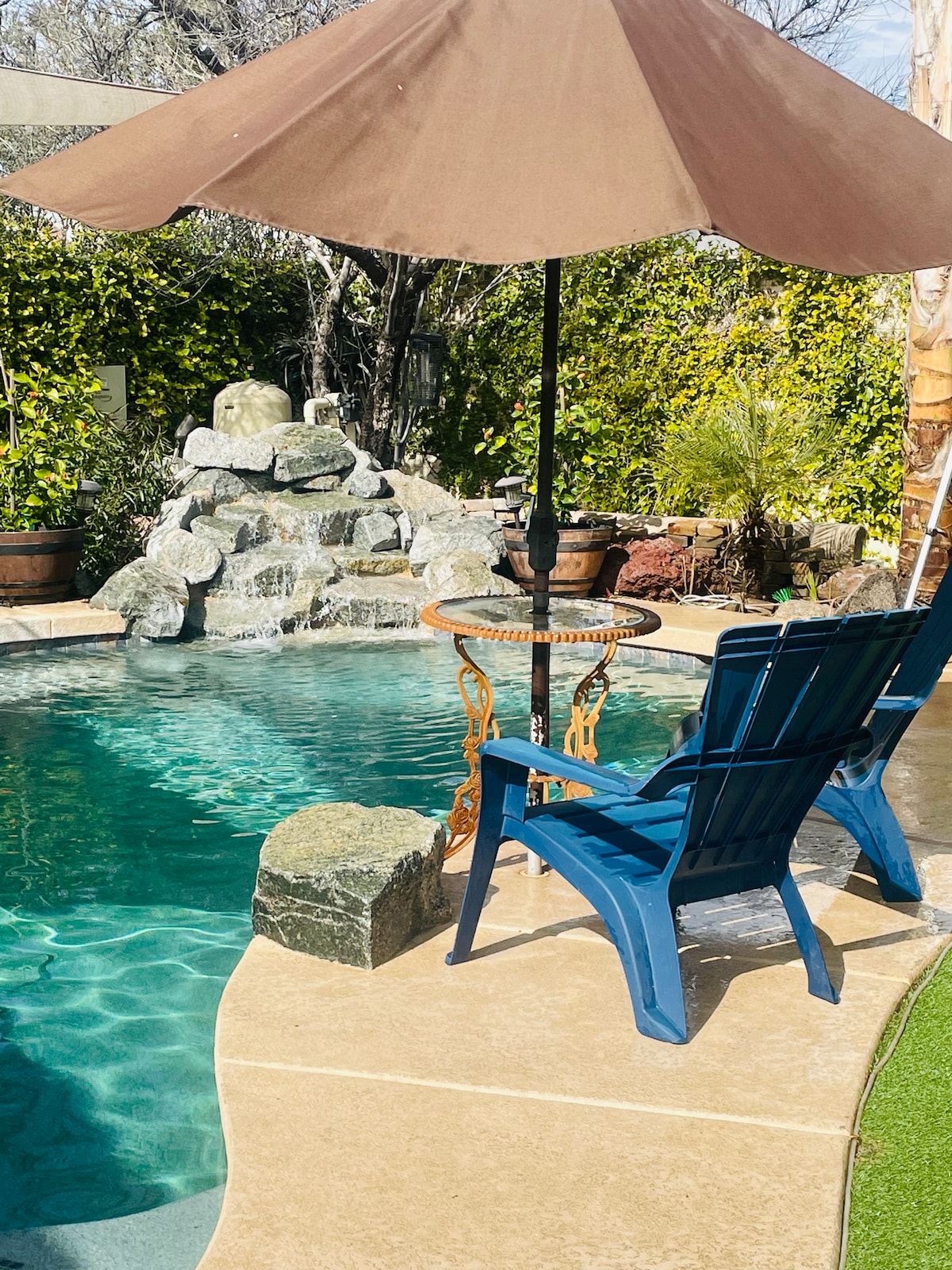 A serene pool area is presented with crystal clear water surrounded by lush greenery. A blue adirondack chair and a small round table are shaded by a brown umbrella, creating a cozy spot for relaxation. A natural stone feature adds texture to the tranquil setting.