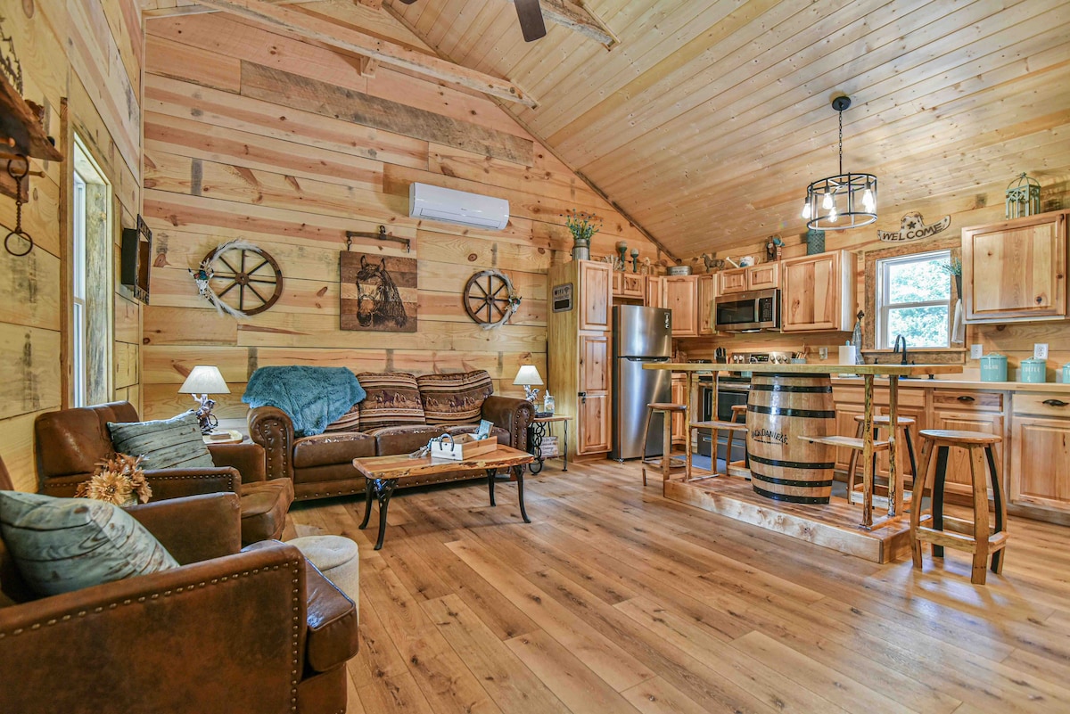 A warm living area features wooden walls and ceiling, complemented by hardwood flooring. A comfortable seating arrangement includes leather sofas positioned near a rustic coffee table. An open kitchen with wooden cabinetry is visible in the background, along with a barrel-style kitchen island.